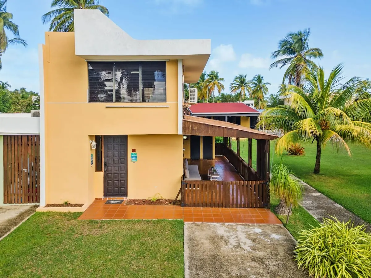 Fachada amarilla de la casa con terraza de madera y palmeras